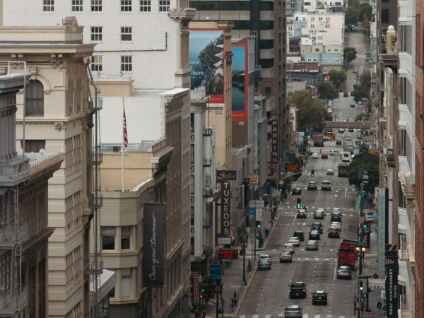 A busy city street in daylight with tall buildings, traffic, pedestrians, and street signs stretching into the distance.