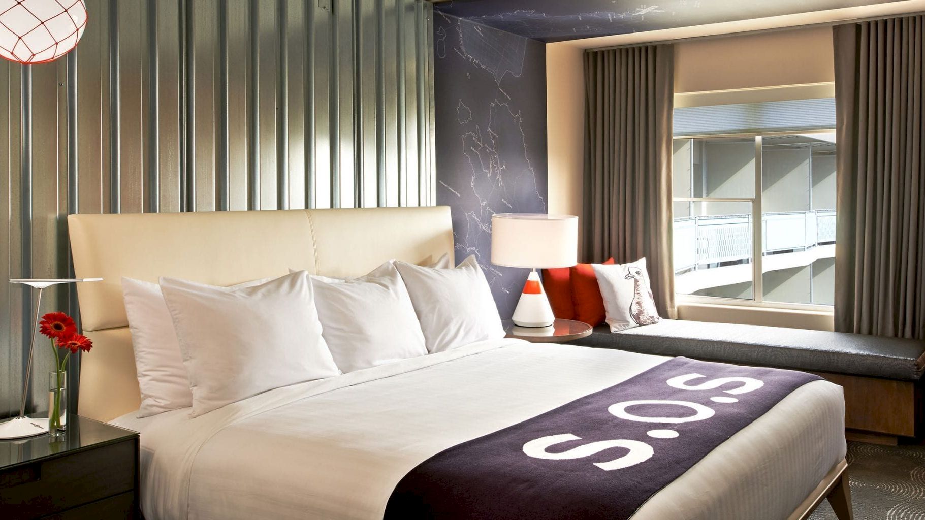 A modern hotel room with a lit bedside lamp, white bed, purple blanket, and map wall decor by tall window blinds.