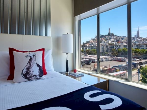 A modern hotel bedroom: a bed with a cat pillow and a city skyline view through large windows.