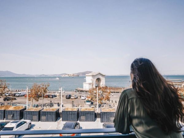 A woman looks out from a car viewpoint over a seaside parking area with cars, an arched building, and distant hills under a clear sky.