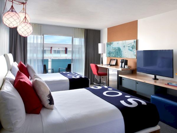 A modern hotel room with twin beds, a TV, a desk, curtains, city/balcony view, and red globe pendant lights.