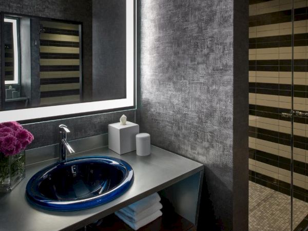 A modern bathroom featuring a blue glass sink, gray textured walls, towels, soap, and a mirror beside the shower glass.