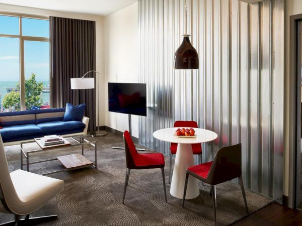 A modern living room and dining area with a blue sofa, flat-screen TV, and a white table with red chairs by tall windows.