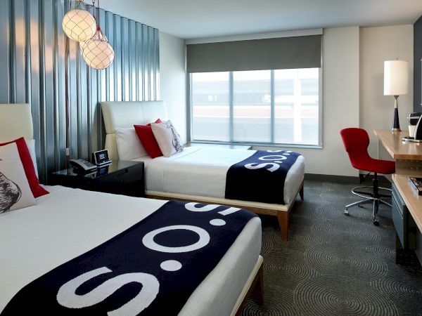A modern hotel room with two beds, red chairs, a desk, large window, and hanging spherical lamps.