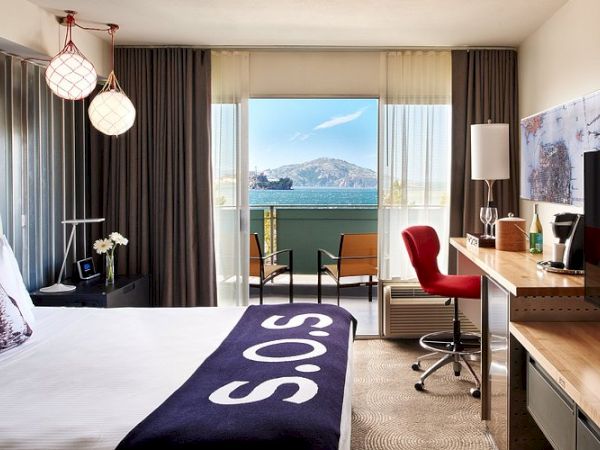 A modern hotel room with a bed, desk, red chair, and a large window showing a lake and mountains.