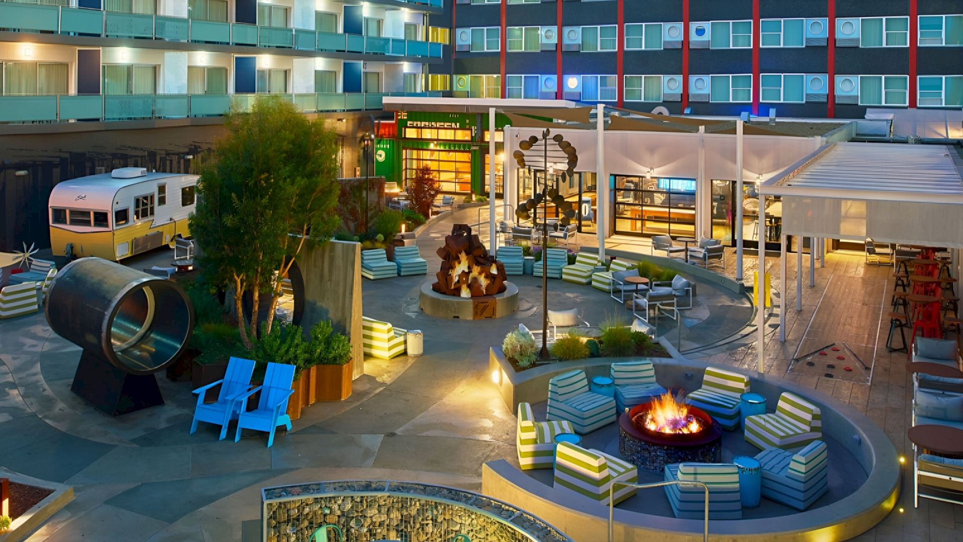 A modern hotel courtyard at night with a fire pit lounge, striped seating, a vintage camper display, and glass doors.