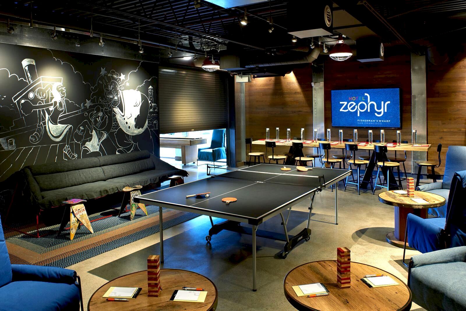 A modern hotel game room with a ping-pong table, sofas, and a &ldquo;Hotel Zephyr&rdquo; sign on the wall.