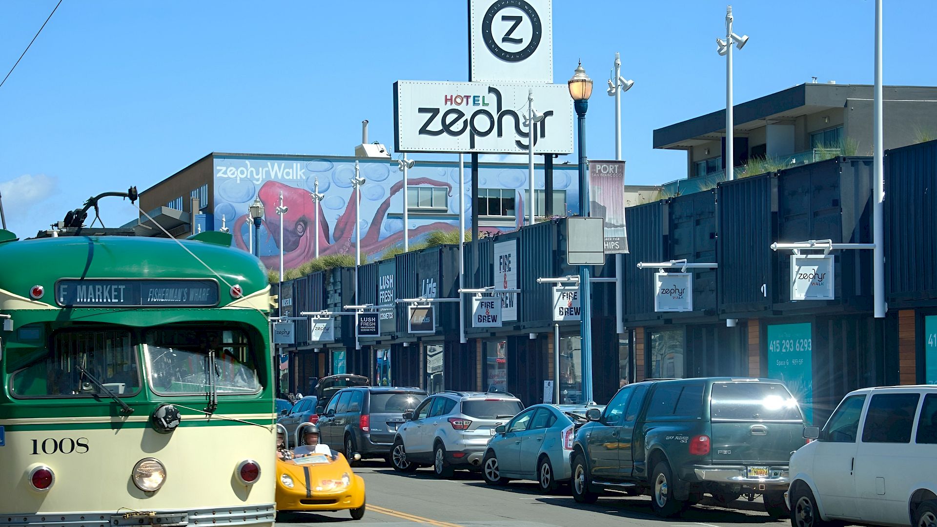 A green vintage trolley and parked cars sit in front of a &ldquo;Hotel Zephyr&rdquo; sign on a sunny street.