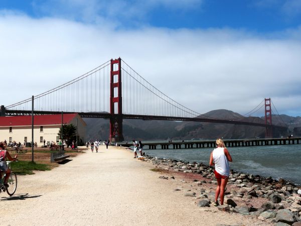 The Golden Gate Bridge looms over a waterfront walkway with visitors, rocks, and boats beneath a bright blue sky.