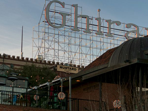 A giant neon-style metal sign with large looping letters rises over brick buildings and greenery in the evening sky.