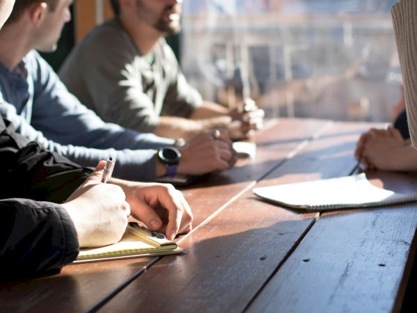 A person writes on a notebook outdoors while others discuss nearby, with pens and papers on a wooden table.