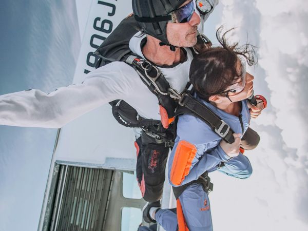 A skydiver in a helmet helps a woman jump from an aircraft, strapped in with safety gear above the clouds.