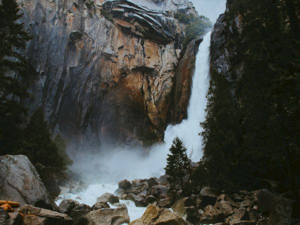 A towering waterfall plunges from a rocky cliff into misty rocks, surrounded by dark pine trees.
