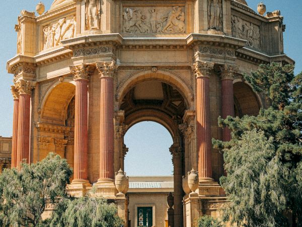 A grand stone monument with towering columns, arched openings, and ornate reliefs and statues carved into its fa&ccedil;ade, standing above trees.