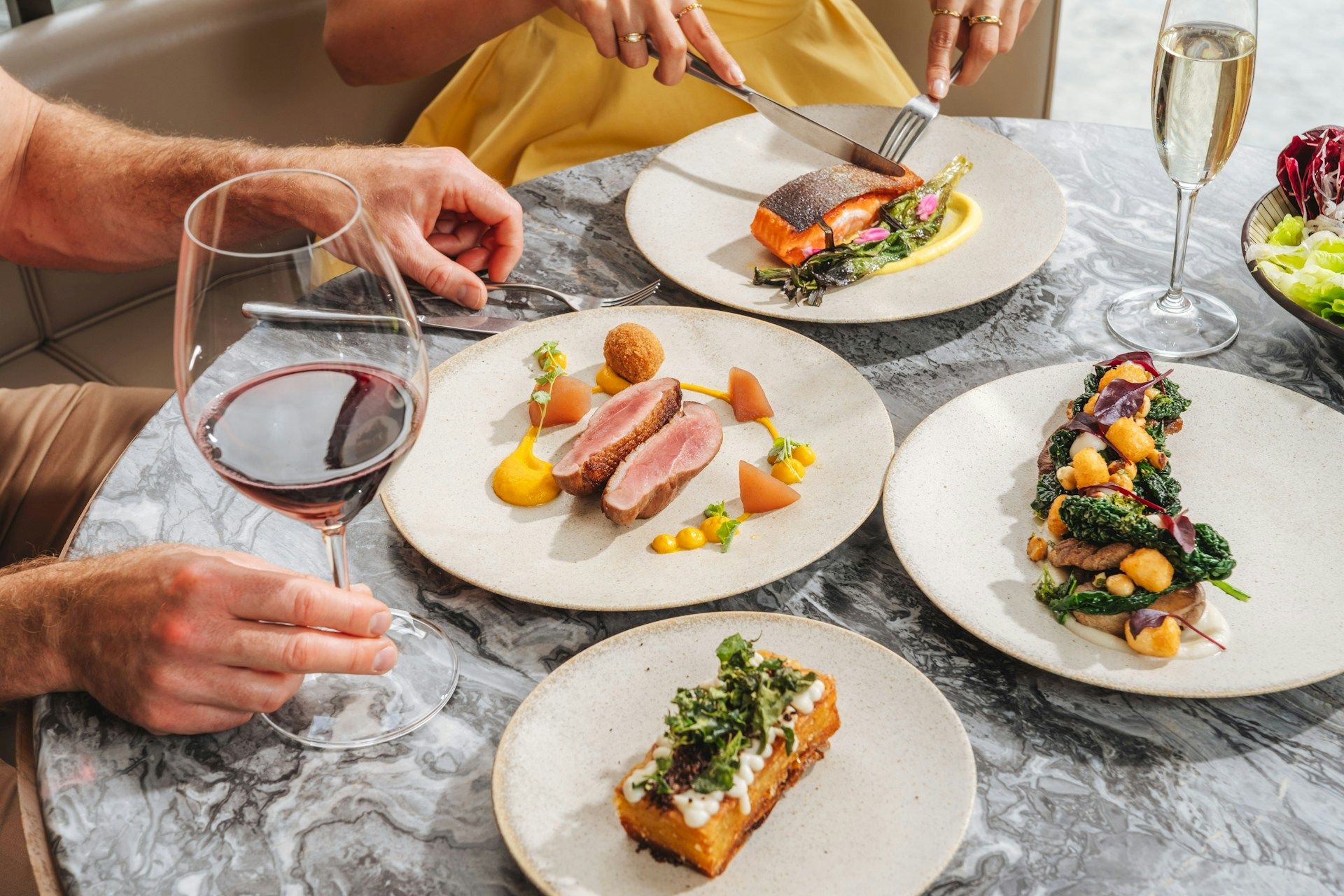 Friends dine as a server plates gourmet entrees with wine on a marble table, sharing a celebratory meal.