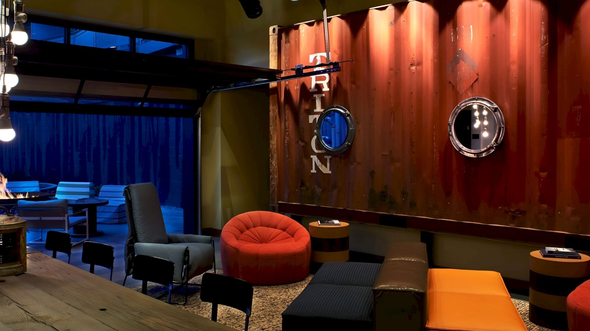 A cozy lounge with orange seating, a fireplace, and an industrial red wall featuring porthole mirrors and lighting.