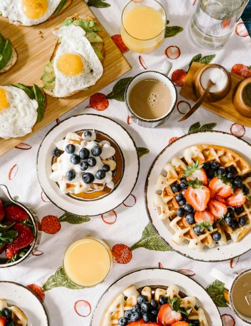 A brunch spread of waffles topped with strawberries and blueberries, fried eggs, and cups of coffee/juice on the table.