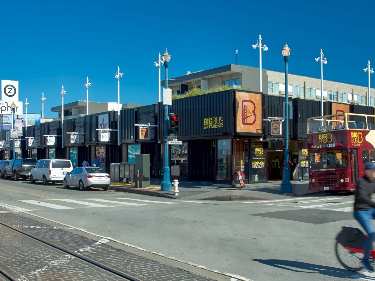 A city street in front of Hotel Zephyr with a Big Bus Tours double-decker, an orange tram, and cars near a container wall.